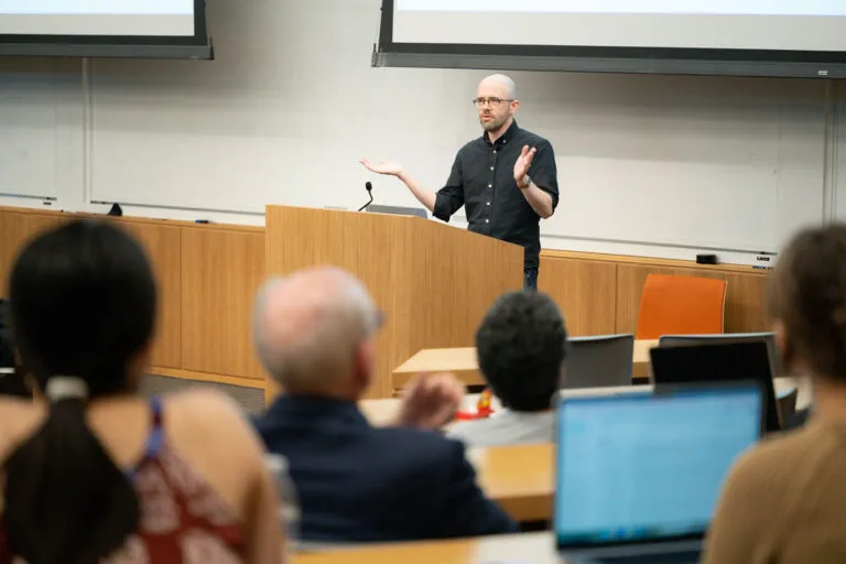 Brian Christian stands at the front of a lecture hall during a talk about AI; students are slightly blurred in the foreground.